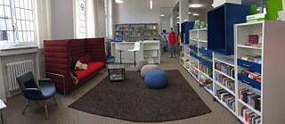 Das Innere einer Bibliothek, mit einem gemütlichen zentralen Raum, einem Sofa, Stühlen und drum herum Bücher in Regalen.